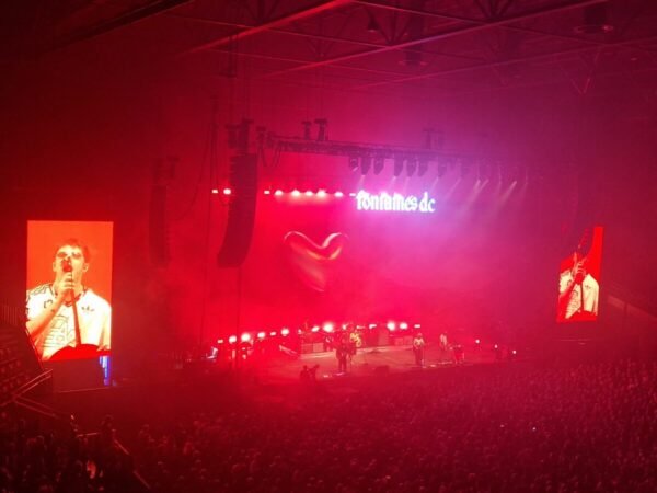 Fontaines D.C. on a red-lit arena stage under a glowing "fontaines dc" sign, with Grian Chatten shown on a side video screen